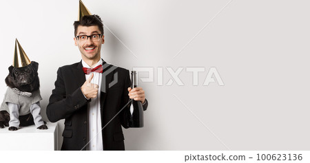 Small black dog wearing party hat and standing near happy man celebrating holiday, owner showing thumb-up and holding champagne bottle, white background Small black dog wearing party hat and standing near happy man celebrating holiday, owner showing thumb-up and holding champagne bottle, white background 100623136