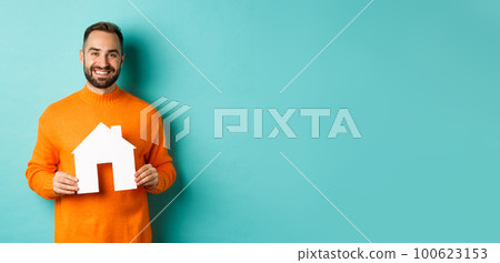 Real estate concept. Happy young man searching for home rent, holding house paper maket and smiling, standing over blue background 100623153