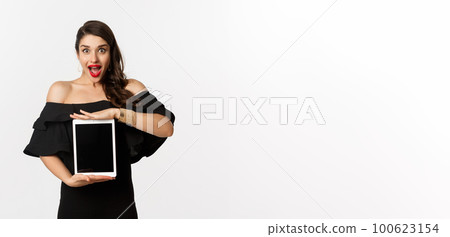 Fashion and shopping concept. Amazed young woman showing online website promo offer on tablet screen, looking excited camera, standing in black dress, white background 100623154