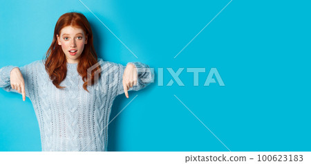 Winter holidays and people concept. Cute teenage girl pointing fingers down, showing advertisement, standing over blue background 100623183
