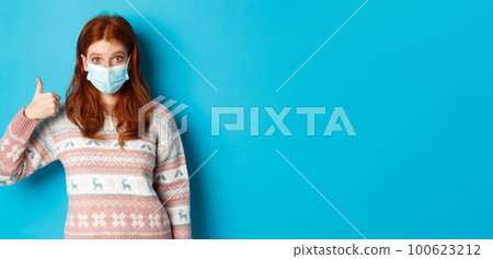 Winter, coronavirus and social distancing concept. Impressed redhead girl in face mask, showing thumb up in approval, like and praise, standing over blue background Winter, coronavirus and social distancing concept. Impressed redhead girl in face mask, showing thumb up in approval, like and praise, standing over blue background 100623212