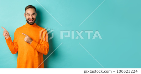 Handsome smiling man pointing fingers left, showing your logo, standing in winter orange sweater, turquoise background Handsome smiling man pointing fingers left, showing your logo, standing in winter orange sweater, turquoise background 100623224