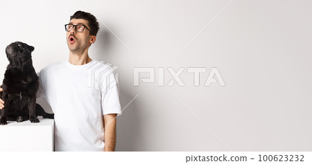Amazed young man in glasses hugging his dog, pet owner and pug staring at upper left corner promo offer, standing over white background 100623232
