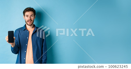 Happy caucasian guy showing empty mobile screen and smiling, standing on blue background 100623245