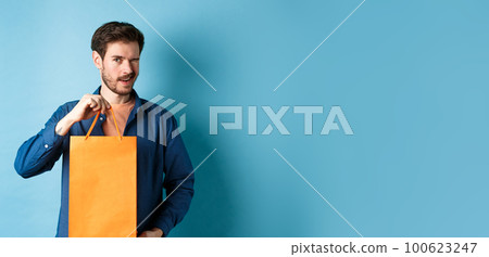 Happy young man showing orange shopping bag and winking, recommending shop discounts, standing on blue background 100623247