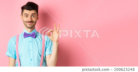 Smiling caucasian man in bow-tie and suspenders showing all okay gesture, recommending something cool, praising promo on pink background 100623248