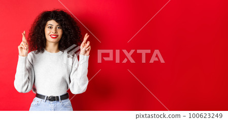 Hopeful young woman with red lips and curly hair, cross fingers for good luck and making wish, praying for dream come true, smiling excited, red background Hopeful young woman with red lips and curly hair, cross fingers for good luck and making wish, praying for dream come true, smiling excited, red background 100623249