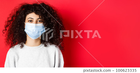 Covid-19 and pandemic concept. Close-up of modern young woman with curly hair, wearing medical mask from coronavirus, smiling at camera, red background 100623335