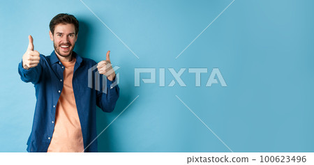 Positive smiling guy showing thumbs up and smiling, complimenting you, praising good job, well done gesture, standing on blue background Positive smiling guy showing thumbs up and smiling, complimenting you, praising good job, well done gesture, standing on blue background 100623496