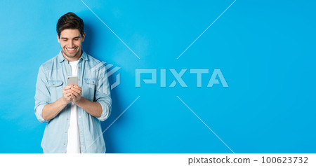 Image of handsome young man using mobile phone, texting on phone and looking pleased, standing over blue background Image of handsome young man using mobile phone, texting on phone and looking pleased, standing over blue background 100623732