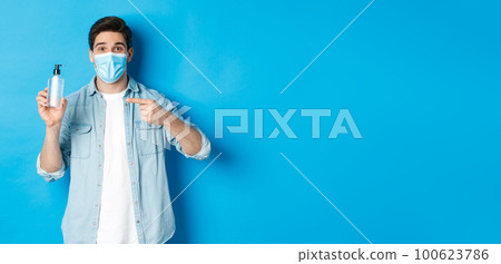 Concept of covid-19, pandemic and social distancing. Handsome guy in medical mask advice to use hand sanitizer, pointing at antiseptic, standing over blue background Concept of covid-19, pandemic and social distancing. Handsome guy in medical mask advice to use hand sanitizer, pointing at antiseptic, standing over blue background 100623786