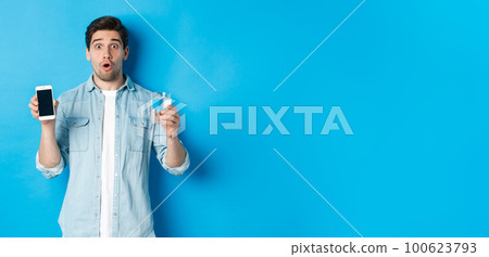 Man looking surprised, showing smartphone screen and glass of water, standing over blue background Man looking surprised, showing smartphone screen and glass of water, standing over blue background 100623793