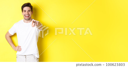 Confident smiling man pointing at you camera, standing in white clothes against yellow background Confident smiling man pointing at you camera, standing in white clothes against yellow background 100623803
