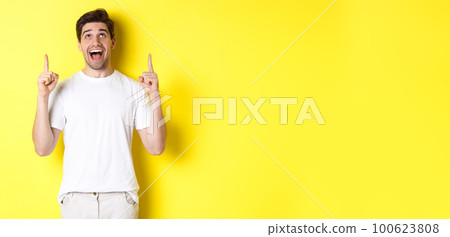 Cheerful young guy in white t-shirt reacting to promo offer, pointing and looking up with amazement, standing over yellow background Cheerful young guy in white t-shirt reacting to promo offer, pointing and looking up with amazement, standing over yellow background 100623808