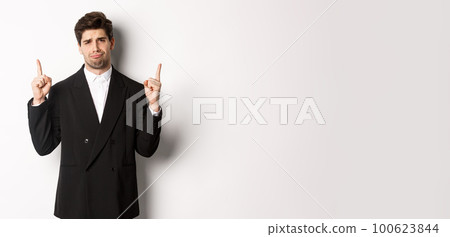 Portrait of reluctant and sad handsome man, complaining and pointing fingers up at something bad, looking jealous, standing over white background 100623844