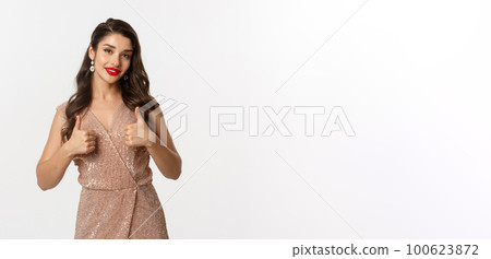 Concept of celebration, holidays and party. Beautiful female model with red lips, wearing glamour dress and showing thumbs up in approval, like and agree, praise good choice 100623872