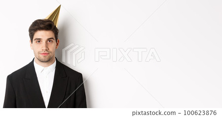 Close-up of handsome man in party hat and trendy suit, celebrating new year, standing against white background 100623876