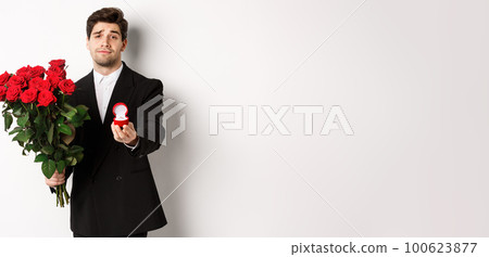 Handsome smiling man in black suit, holding roses and engagement ring, making a proposal to marry him, standing against white background Handsome smiling man in black suit, holding roses and engagement ring, making a proposal to marry him, standing against white background 100623877