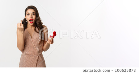 Excited young woman in glamour dress being engaged, showing finger with ring and looking happy, receive marriage proposal, standing over white background Excited young woman in glamour dress being engaged, showing finger with ring and looking happy, receive marriage proposal, standing over white background 100623878