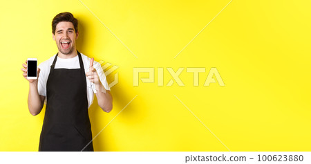 Happy waiter showing mobile screen and thumb up, recommending cafe restaurant app, standing over yellow background Happy waiter showing mobile screen and thumb up, recommending cafe restaurant app, standing over yellow background 100623880