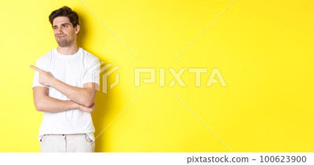 Unimpressed young man complaining, pointing and looking with skeptical face at bad product, standing against yellow background 100623900