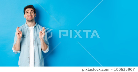 Lucky guy praying and making wish with crossed fingers, looking up with pleading face, standing against blue background Lucky guy praying and making wish with crossed fingers, looking up with pleading face, standing against blue background 100623907