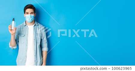 Concept of covid-19, pandemic and social distancing. Young man in medical mask showing hand sanitizer, standing against blue background Concept of covid-19, pandemic and social distancing. Young man in medical mask showing hand sanitizer, standing against blue background 100623965