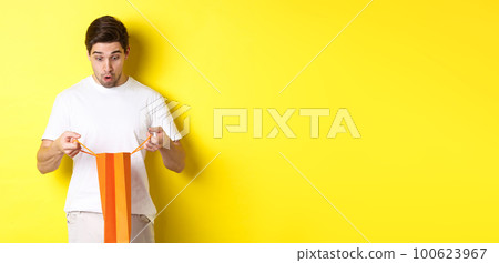 Surprised boyfriend open bag with gift, looking inside amazed, standing over yellow background 100623967