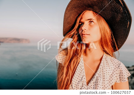 Portrait of happy young woman wearing summer black hat with large brim at beach on sunset. Closeup face of attractive girl with black straw hat. Happy young woman smiling and looking at camera at sea. 100627037