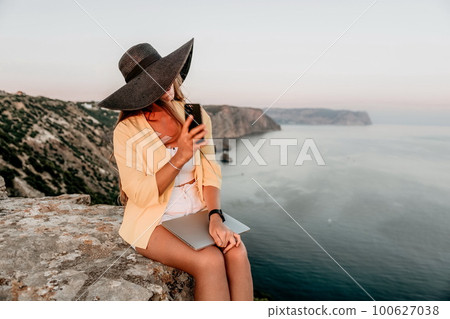 Digital nomad, Business woman working on laptop by the sea. Pretty lady typing on computer by the sea at sunset, makes a business transaction online from a distance. Freelance, remote work on vacation 100627038