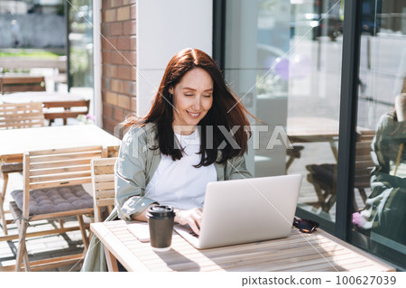 Adult smiling brunette woman forty years in stylish shirt working on laptop in cafe at city street 100627039