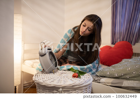 A sad woman looks at her white alarm clock on her bedside table. 100632449