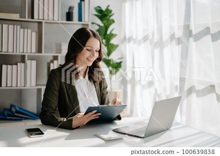 Confident business expert attractive smiling young woman typing laptop ang holding digital tablet on desk in creative home office. Confident business expert attractive smiling young woman typing laptop ang holding digital tablet on desk in creative home office. 100639838