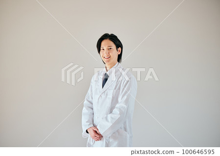 A young man in a white coat standing with his hands folded in front 100646595