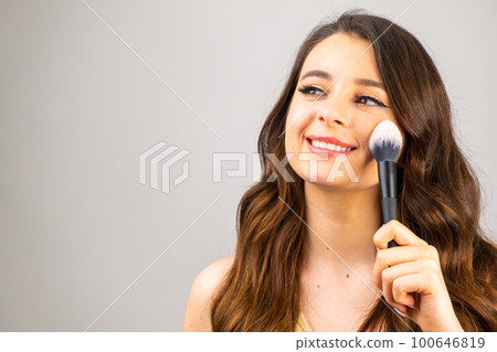 Portrait of an attractive lovely young woman holding makeup blusher brush on the grey background with copy space. Portrait of an attractive lovely young woman holding makeup blusher brush on the grey background with copy space. 100646819
