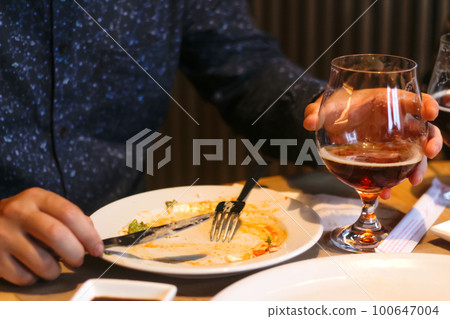 After meal concept. Dirty dishes from the tables in restaurant. Empty dirty plate after meal with fork inside next to napkins with chopsticks, male hand. Man holding glass with beer or whiskey 100647004