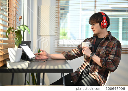Satisfied millennial man freelancer in wireless headphone using laptop computer on black wooden desk at home office 100647906