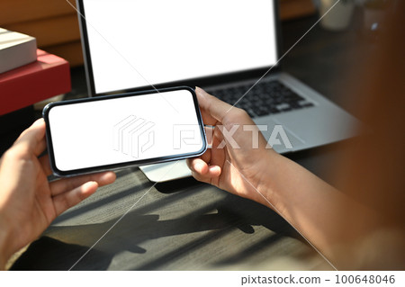 Mockup image of woman hands using smart phone at working desk front of laptop computer. Blank screen for advertising text Mockup image of woman hands using smart phone at working desk front of laptop computer. Blank screen for advertising text 100648046