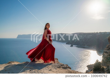 A woman in a red flying dress fluttering in the wind, against the backdrop of the sea. 100649742