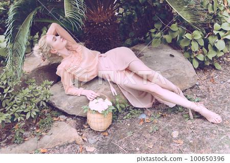 Sensual young blonde beautiful cute and elegant woman wearing pink sitting on the rock and looking into the camera over tropical green jungle palm background in China 100650396