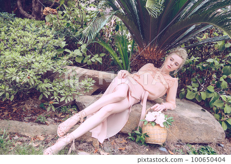Sensual young blonde beautiful cute and elegant woman wearing pink sitting on the rock and looking into the camera over tropical green jungle palm background in China Sensual young blonde beautiful cute and elegant woman wearing pink sitting on the rock and looking into the camera over tropical green jungle palm background in China 100650404