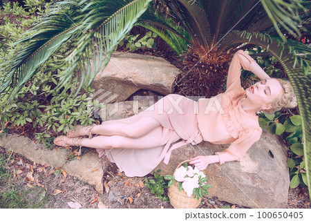 Sensual young blonde beautiful cute and elegant woman wearing pink sitting on the rock and looking into the camera over tropical green jungle palm background in China 100650405