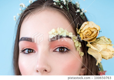 Cosmetics and manicure. Close-up portrait of attractive woman with dry flowers on her face and hair, pastel color, perfect make-up and skin on blue background. Fresh, trendy, spring retouched portrait Cosmetics and manicure. Close-up portrait of attractive woman with dry flowers on her face and hair, pastel color, perfect make-up and skin on blue background. Fresh, trendy, spring retouched portrait 100650763