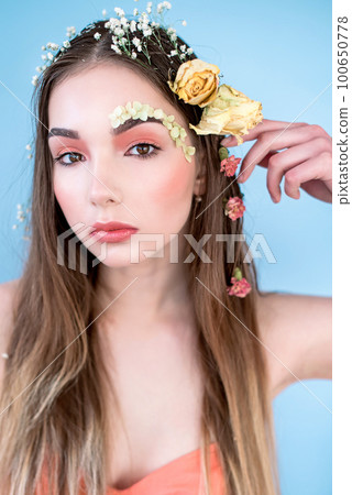 Cosmetics and manicure. Close-up portrait of attractive woman with dry flowers on her face and hair, pastel color, perfect make-up and skin on blue background. Fresh, trendy, spring retouched portrait 100650778