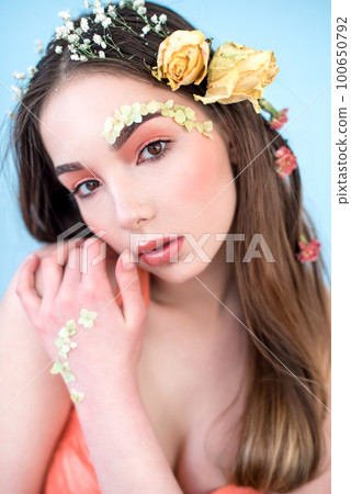 Cosmetics and manicure. Close-up portrait of attractive woman with dry flowers on her face and hair, pastel color, perfect make-up and skin on blue background. Fresh, trendy, spring retouched portrait 100650792