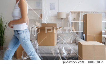 Home moving. Flat relocation. Removal service. Unrecognizable woman leaving room with carton boxes in modern apartment with polyethylene covered furniture. 100653034