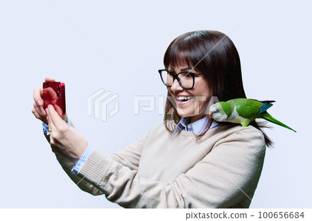 Happy woman with parrot on shoulder taking selfie on smartphone, white background 100656684
