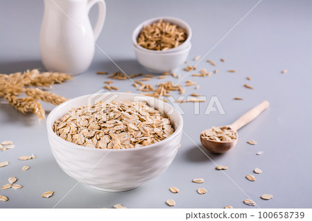 Raw oatmeal in a bowl with a wooden spoon and ears on a gray background. 100658739