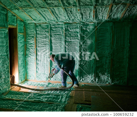 Male builder insulating wooden frame house. Man worker spraying polyurethane foam inside of future cottage, using plural component gun. Construction and insulation concept. 100659232