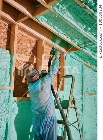 Male builder insulating wooden frame house. Man worker spraying polyurethane foam inside of future cottage, standing on ladder, using plural component gun. Construction and insulation concept. 100659234